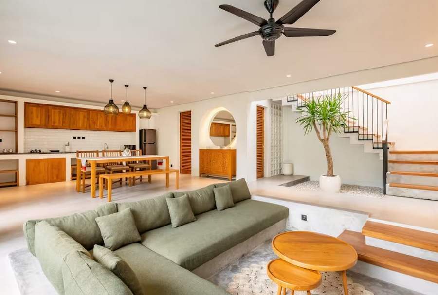 This image shows the living area of a Balinese villa that has a sunken seating area, a couch with natural fiber green cusions, and teakwood furniture including tables and cabinet doors in the kitchen.