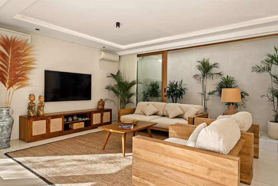 This image shows a modern Bali villa living room that features teak wood furniture with cream colored cushions, a reclaimed teak wood TV stand with wicker doors, a woven authentic area rug, and large glass sliding doors that open up to a small exterior garden.