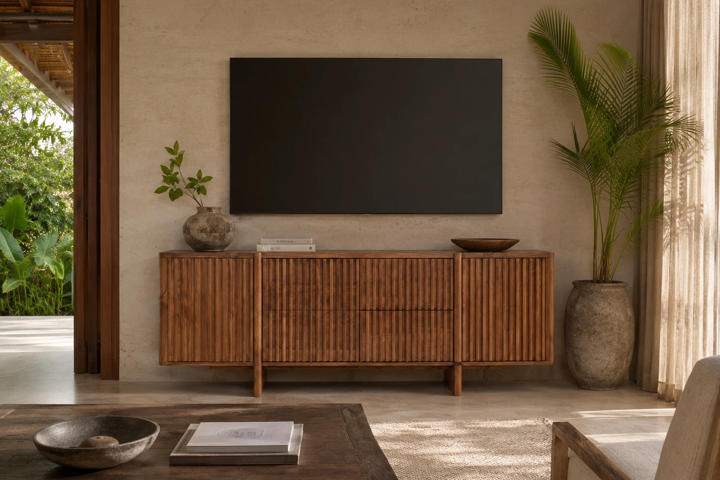 A slatted teak wood media console sits beneath a wall-mounted TV in a luxury, sunlit living room featuring tropical plants and a view of a lush garden.