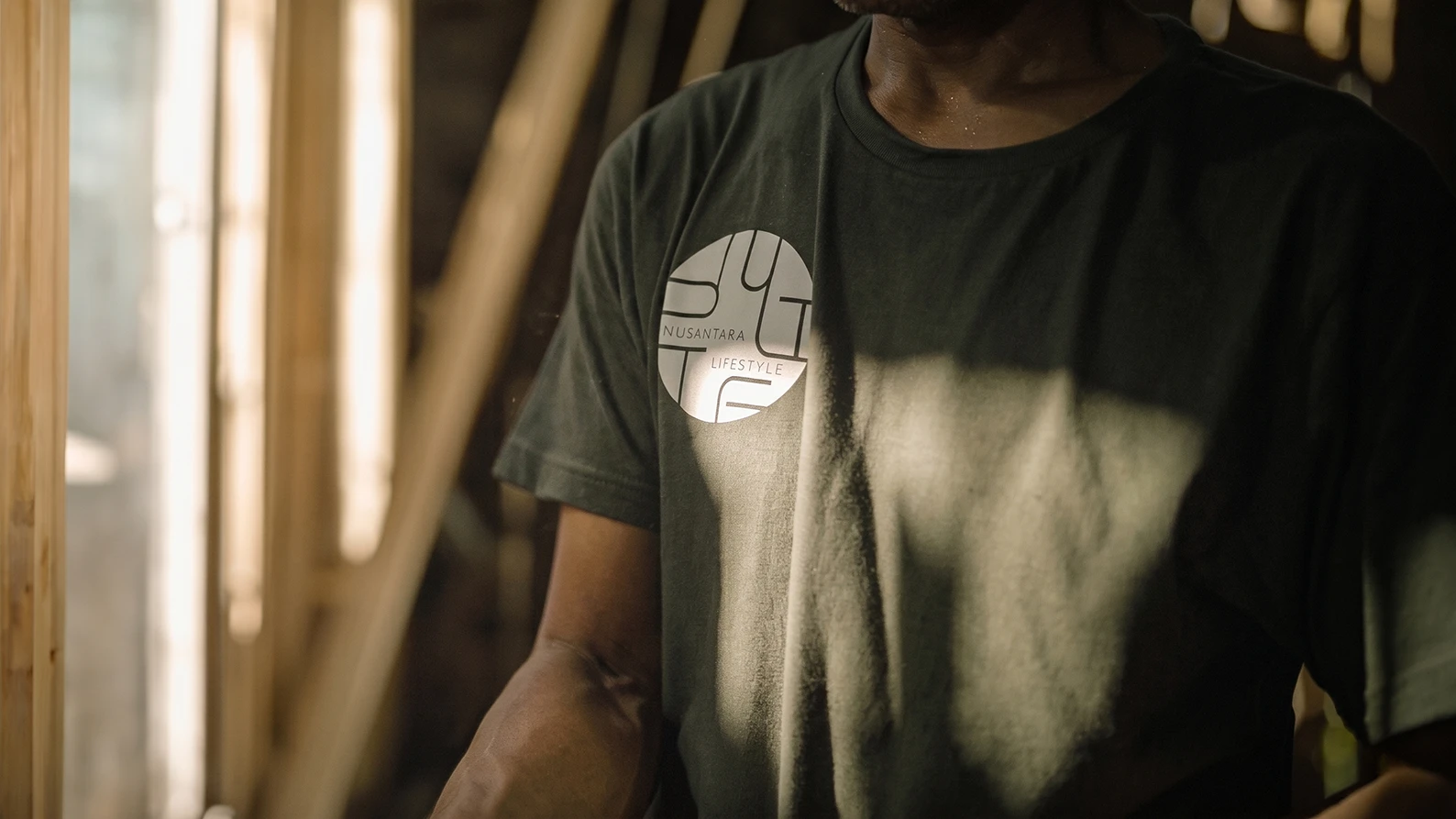 Close-up of a worker in a carpentry workshop wearing a dark t-shirt featuring the white Nusantara Lifestyle logo.