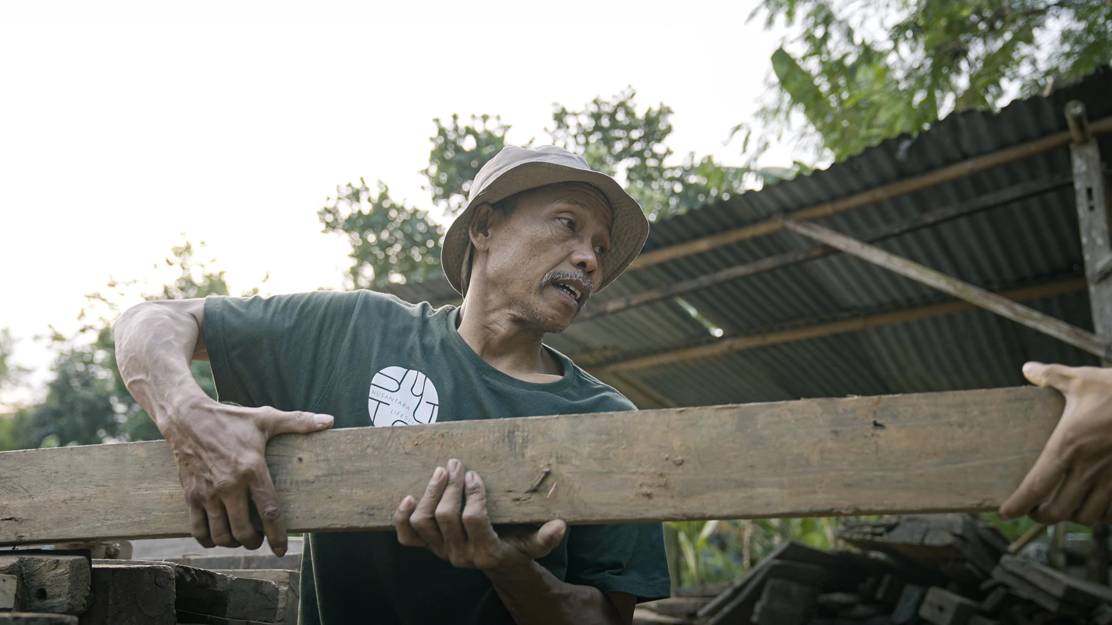 A Nusantara Lifestyle worker handling a reclaimed teak plank during material recovery, showing the timber's original character before manufacturing.